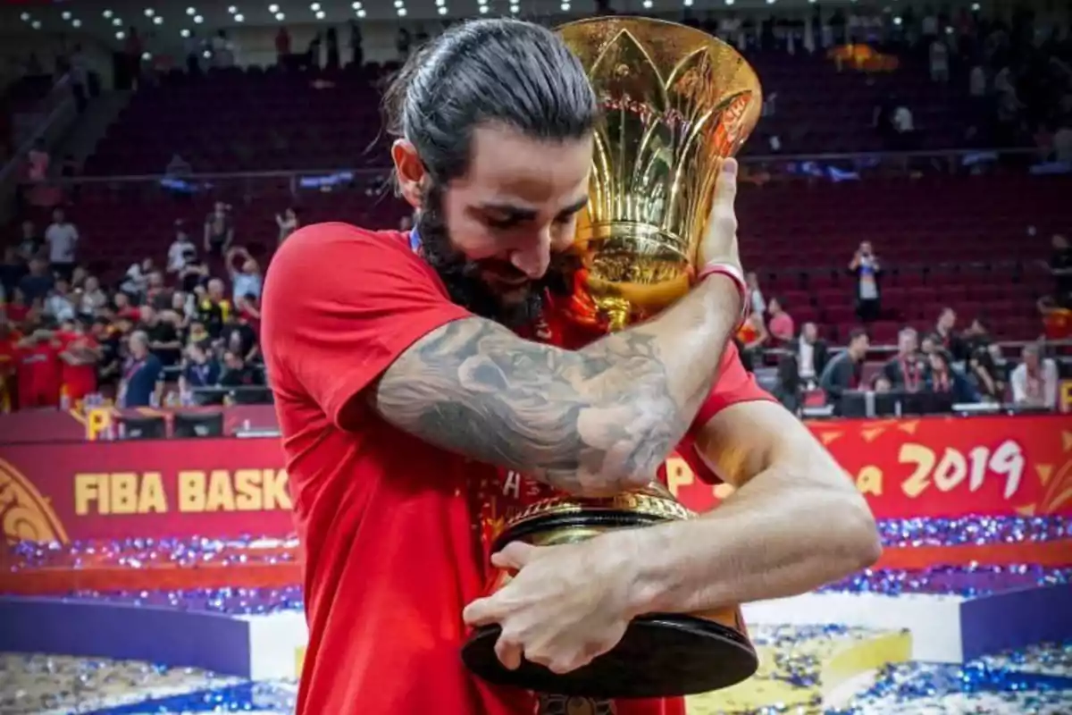 Jugador de baloncesto abrazando un trofeo dorado en una cancha con público y confeti celebrando una victoria