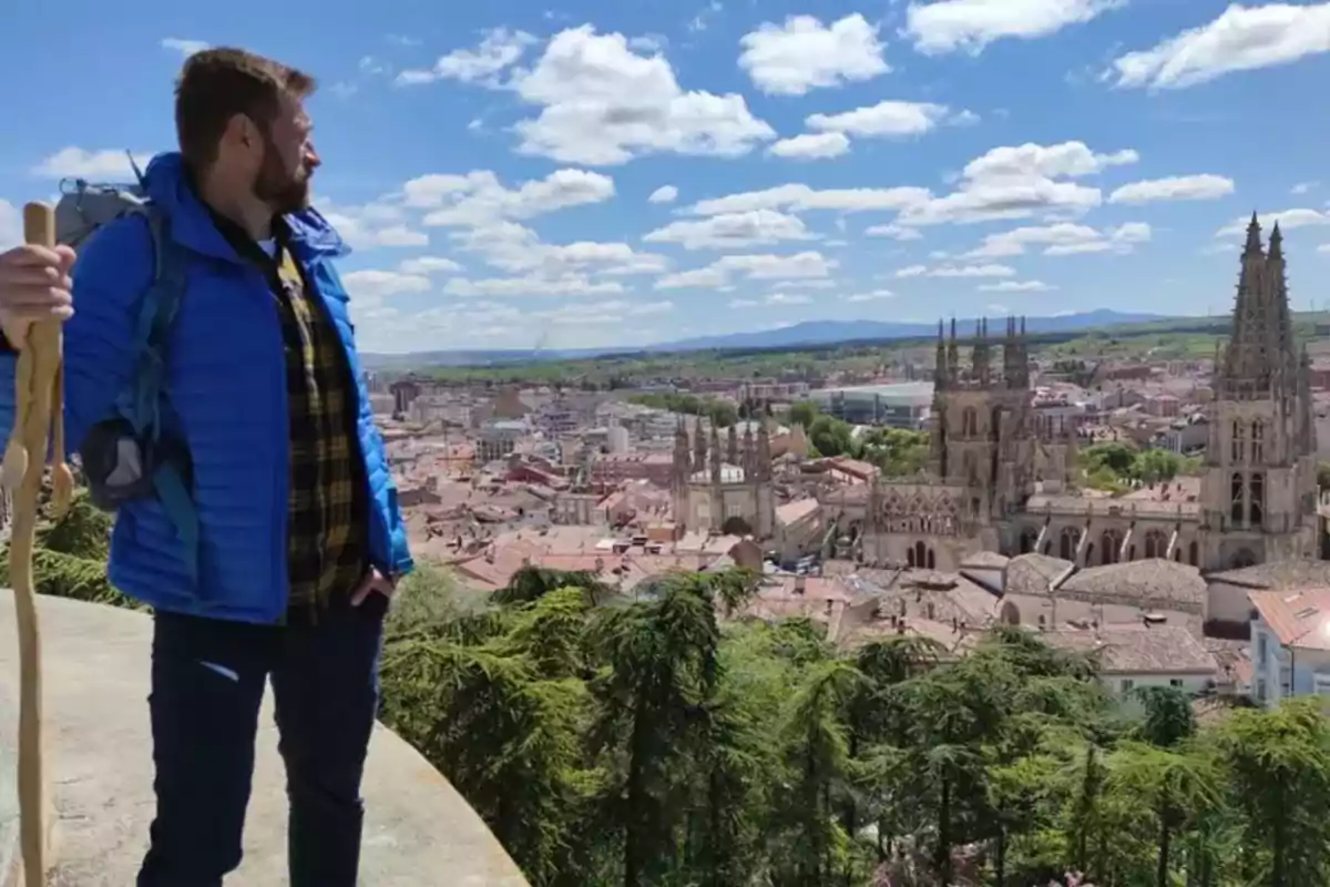 Persona con chaqueta azul y bastón de senderismo observando una ciudad con una catedral gótica al fondo en un día soleado.