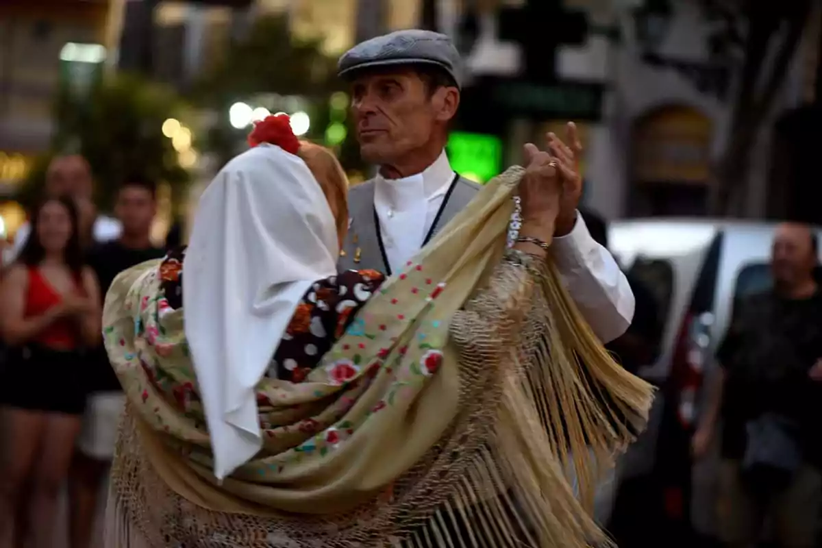 Una pareja de personas mayores baila en una calle, él lleva una boina gris y ella un pañuelo blanco en la cabeza, ambos visten ropa tradicional.