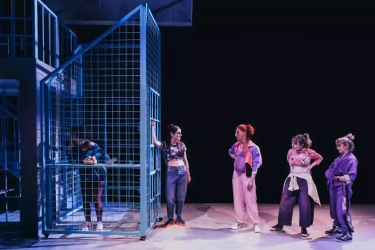 Un grupo de mujeres vestidas con ropa deportiva se encuentra en un escenario, una de ellas está dentro de una estructura metálica mientras las otras cuatro están afuera observándola.