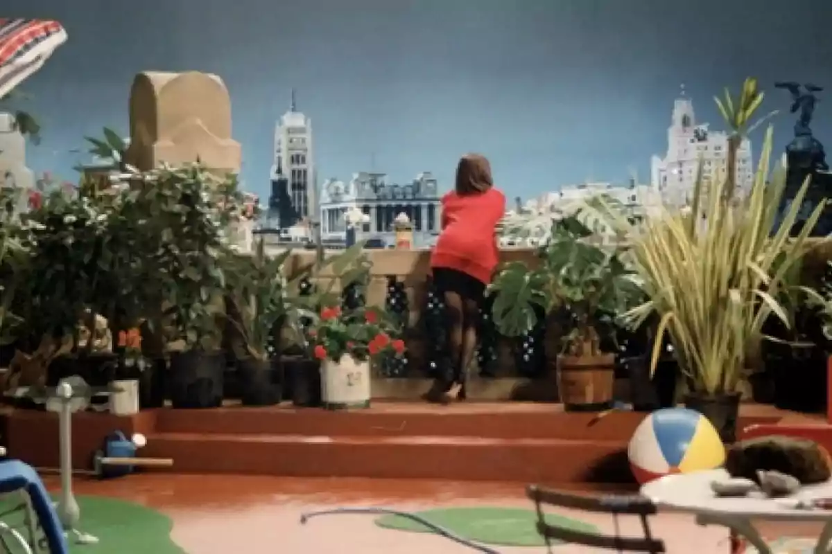 Una persona con un suéter rojo se apoya en una barandilla en una terraza llena de plantas, con una vista de la ciudad al fondo.