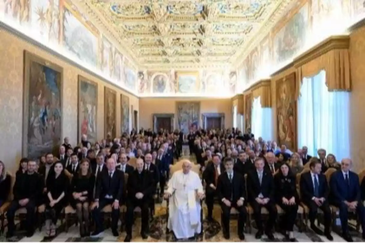 Un grupo de personas sentadas en una sala decorada con un techo ornamentado y pinturas en las paredes.