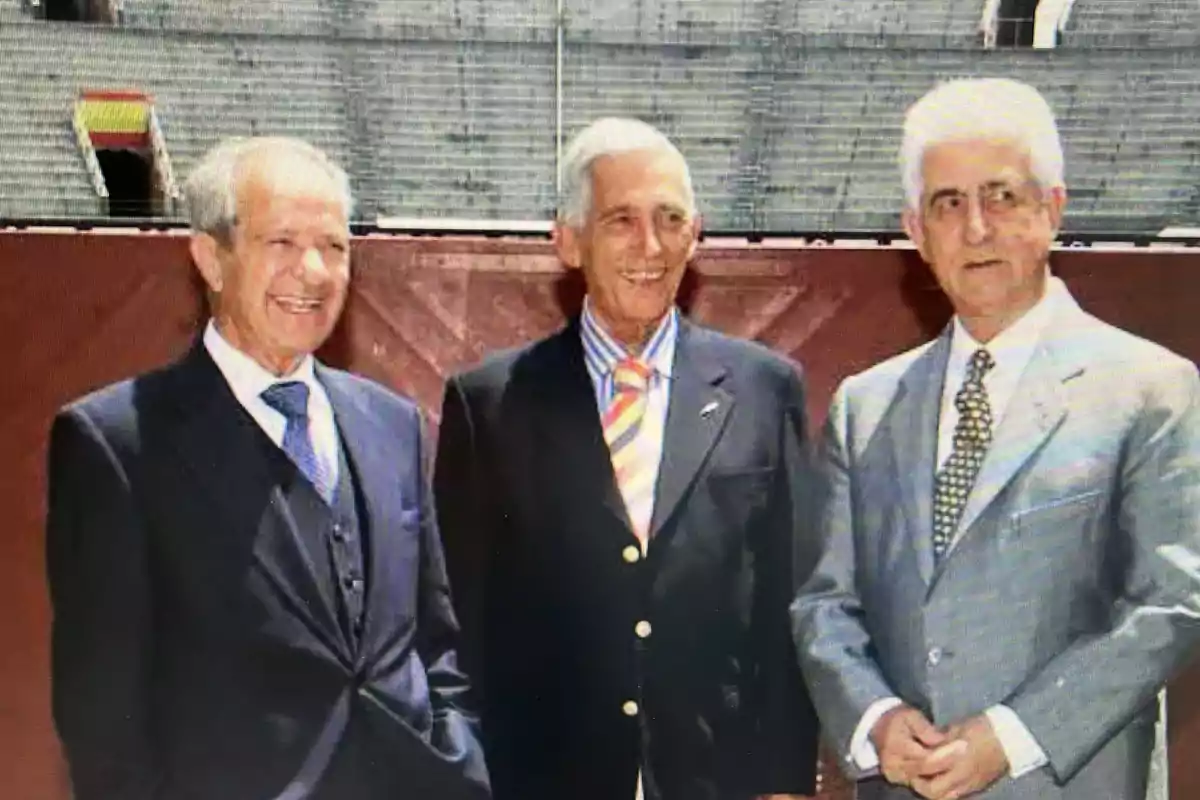 Tres hombres mayores vestidos con trajes formales posan juntos y sonríen frente a una estructura de fondo.