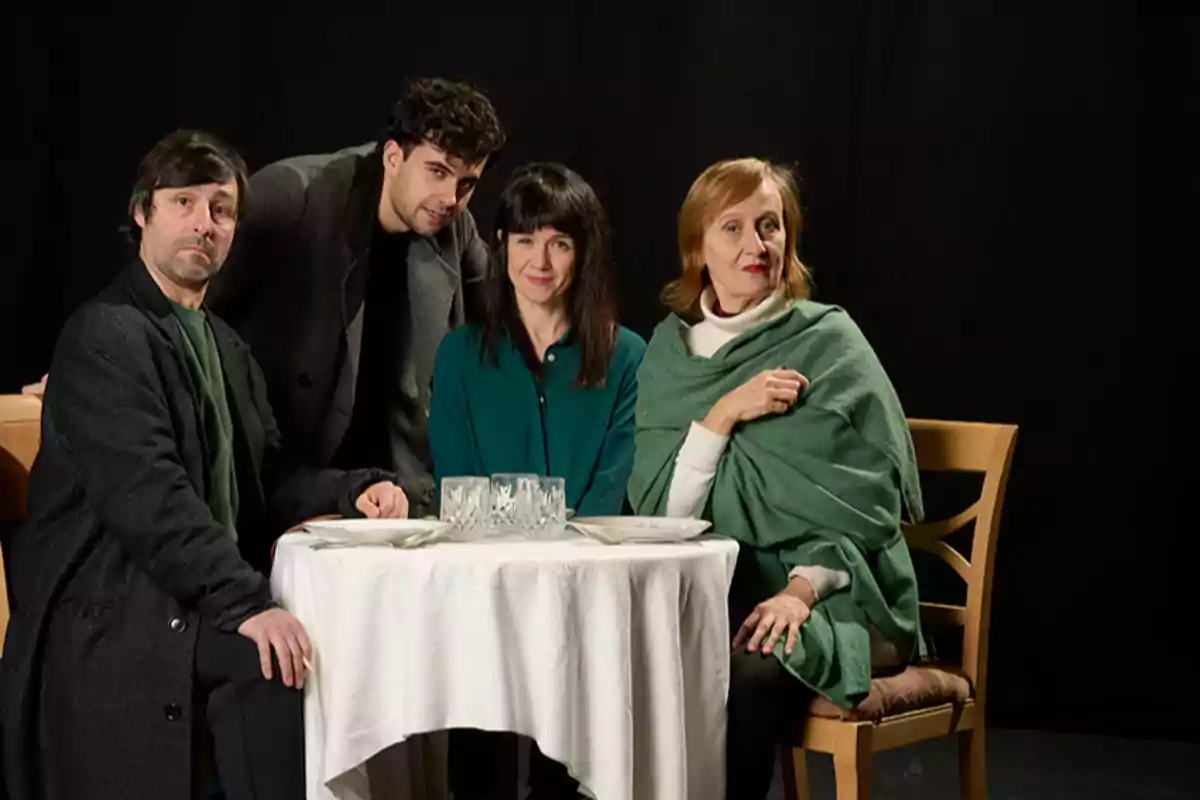 Un grupo de cuatro personas posando alrededor de una mesa redonda con un mantel blanco y copas de cristal, en un ambiente con fondo oscuro.