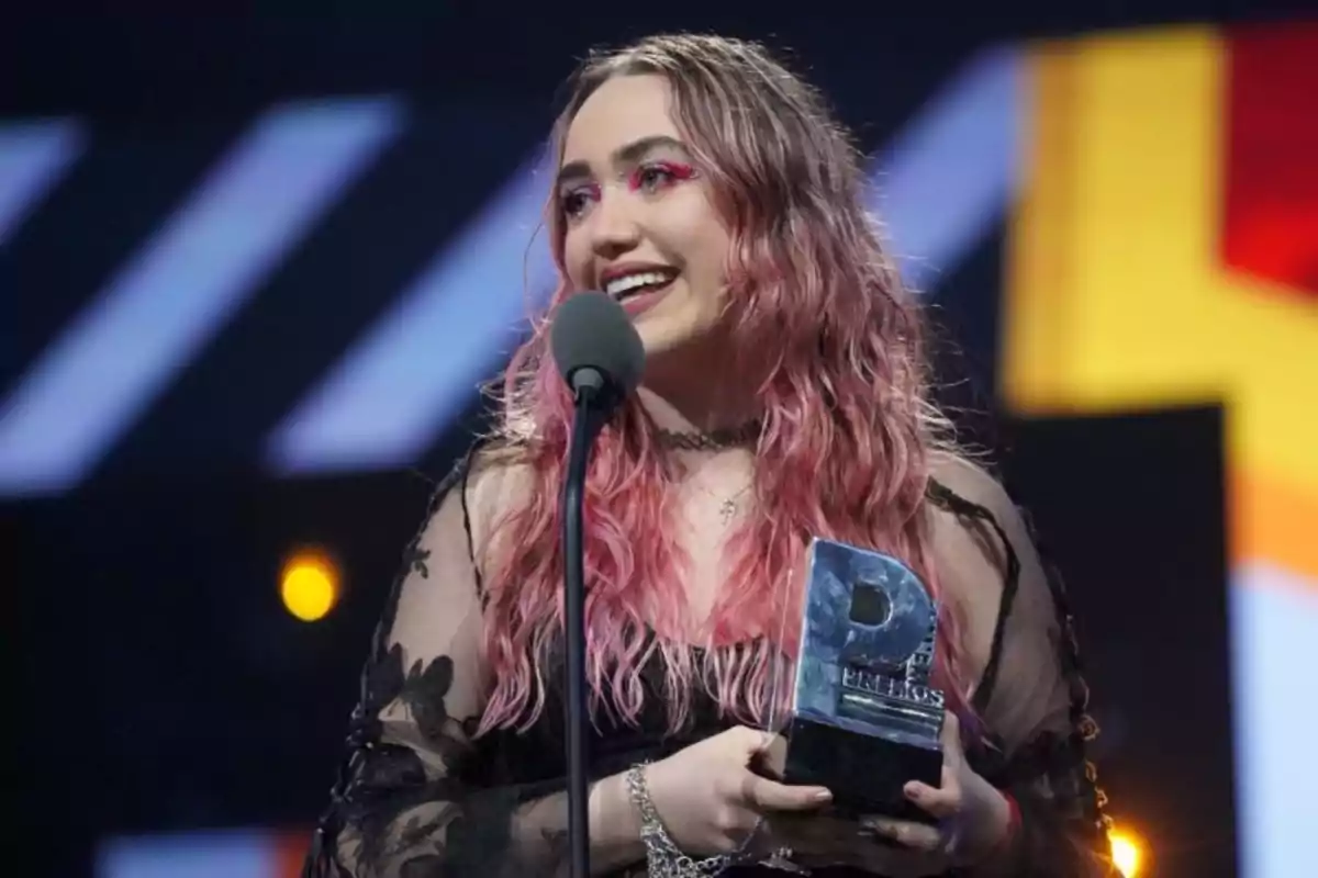 Una persona con cabello rosado y maquillaje brillante sostiene un trofeo mientras habla en un micrófono en un escenario iluminado.