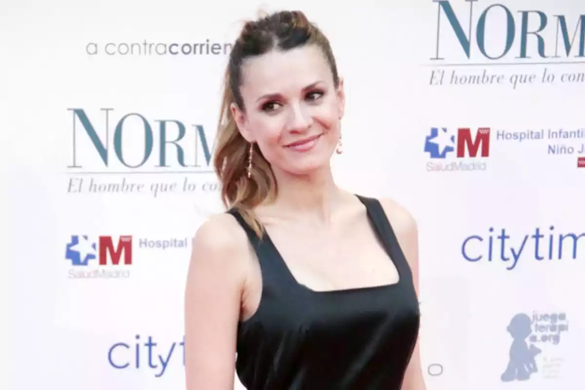 Una persona posando en un evento con un vestido negro y fondo de logotipos. Una persona posando en un evento con un vestido negro y fondo de logotipos.