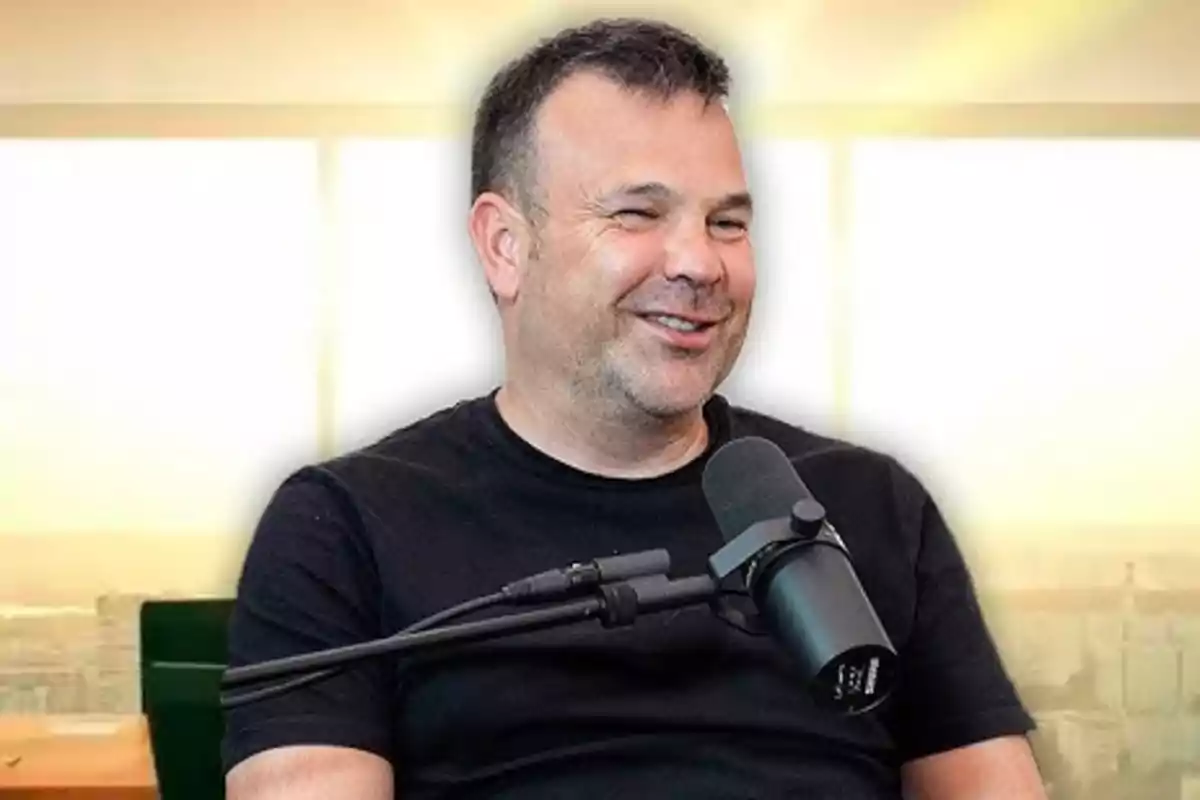 Un hombre sonriente con camiseta negra frente a un micrófono en un estudio iluminado.