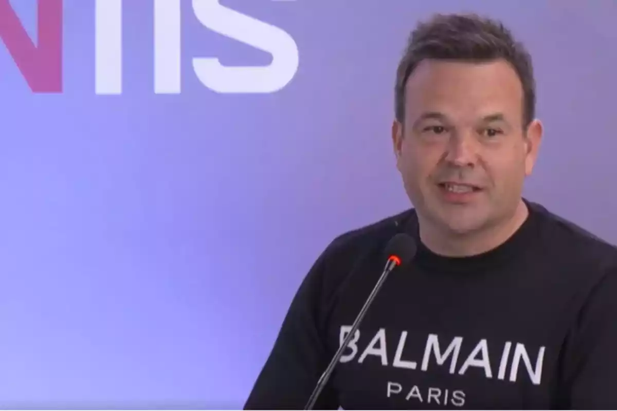 Un hombre con una camiseta de Balmain Paris habla frente a un micrófono en un entorno con fondo azul y letras blancas y rojas.