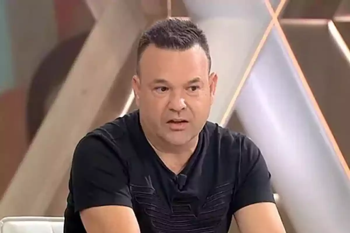 Un hombre con camiseta negra está sentado en un estudio de televisión.