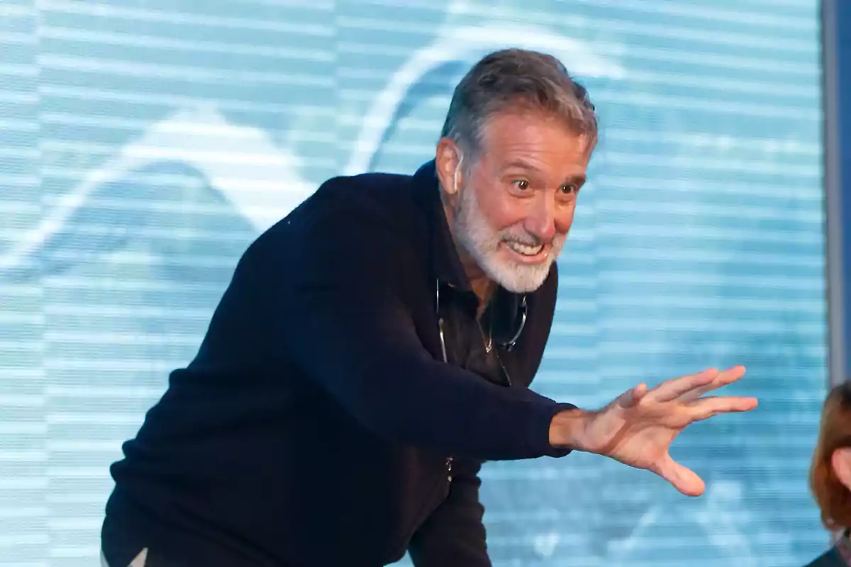 Un hombre con barba y cabello canoso gesticula con entusiasmo frente a un fondo azul.