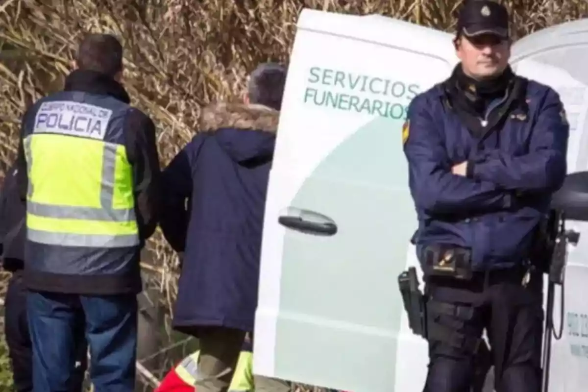 Un policía de pie junto a una furgoneta de servicios funerarios mientras otras personas están de espaldas.