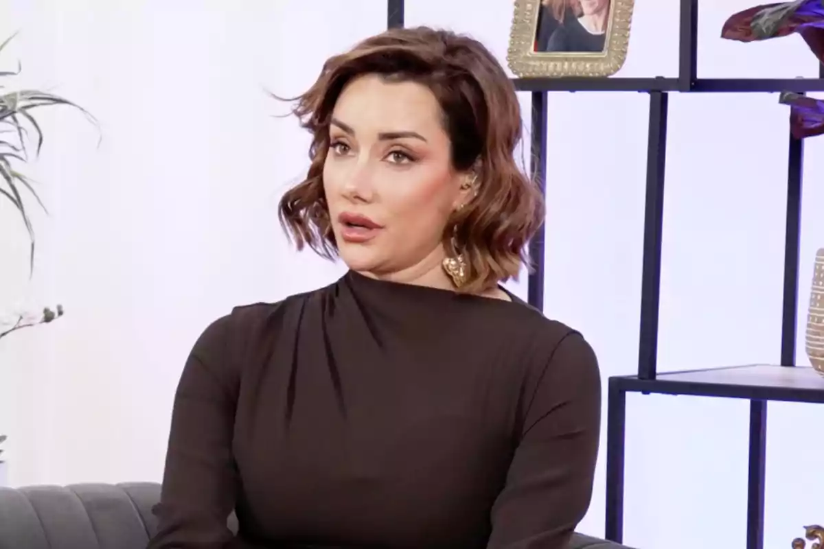 Adara Molinero, vistiendo un vestido oscuro, está sentada en un sofá frente a un fondo con estantes y plantas. Captura de 'En las mejores familias' Adara Molinero, vistiendo un vestido oscuro, está sentada en un sofá frente a un fondo con estantes y plantas. Captura de 'En las mejores familias'