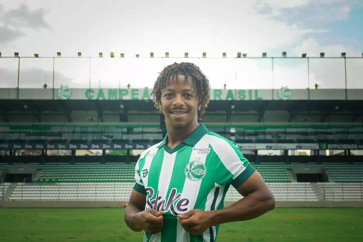 Un jugador de fútbol sonríe mientras sostiene la camiseta de su equipo en un estadio vacío. Un jugador de fútbol sonríe mientras sostiene la camiseta de su equipo en un estadio vacío.