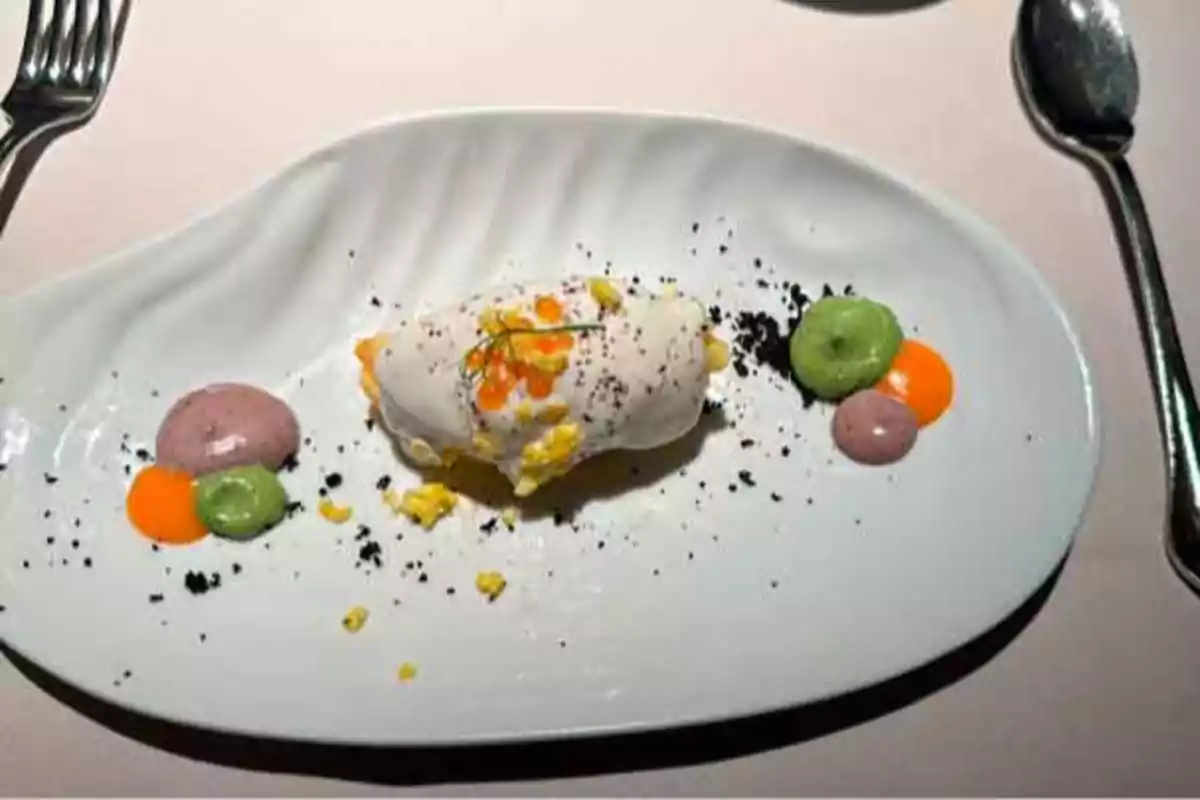 Un plato gourmet con una presentación artística que incluye una porción de comida cubierta con una salsa blanca y decorada con pequeñas porciones de puré de colores y migas negras, acompañado de utensilios de mesa.