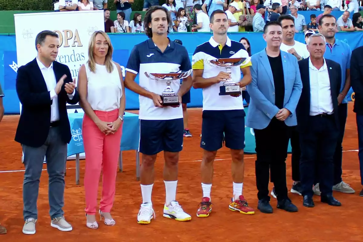 Entrega de premios en un torneo de tenis, con los ganadores sosteniendo trofeos y acompañados por varias personas en una cancha de arcilla.