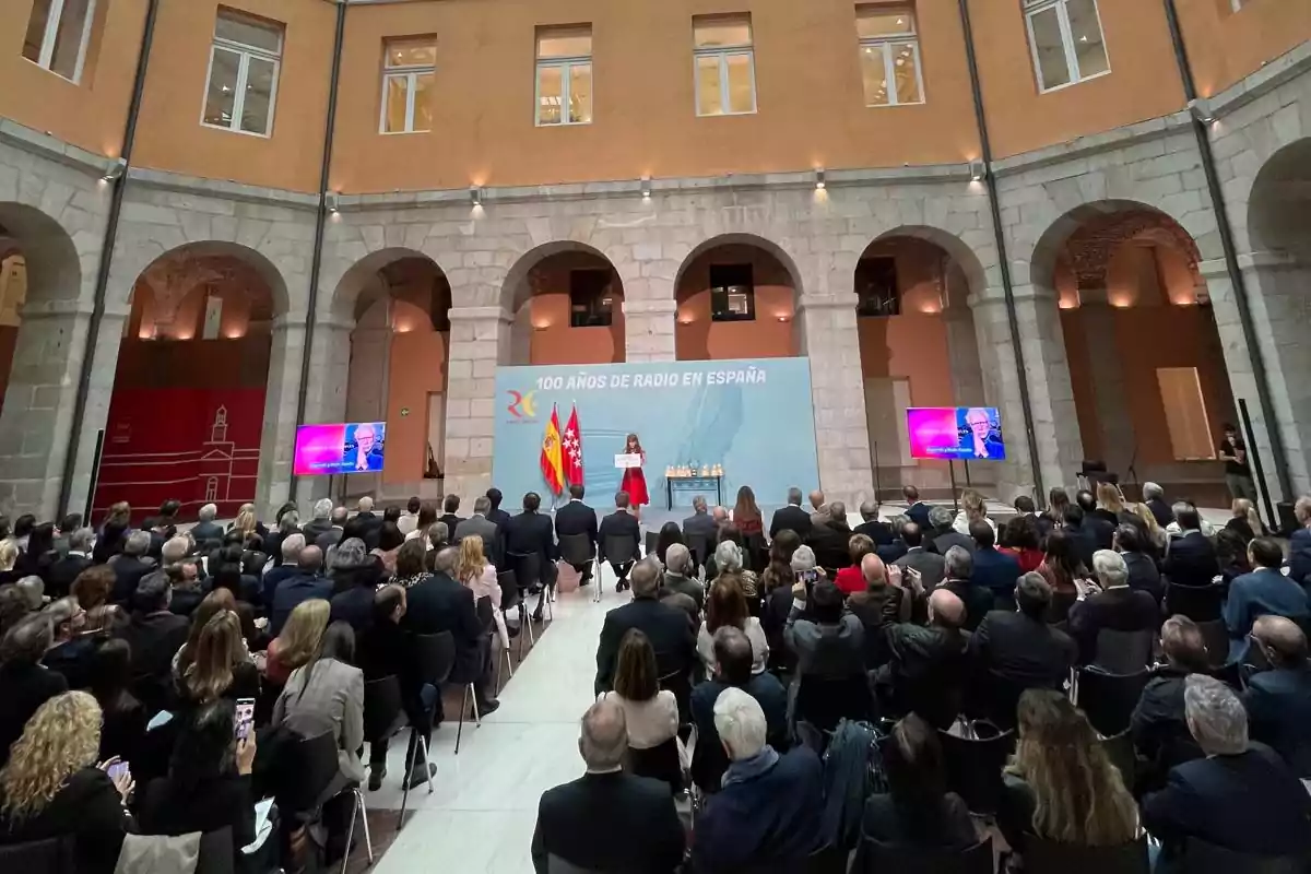 Una multitud asiste a un evento en un edificio histórico con arcos y una pantalla que muestra una presentación sobre los 100 años de radio en España.