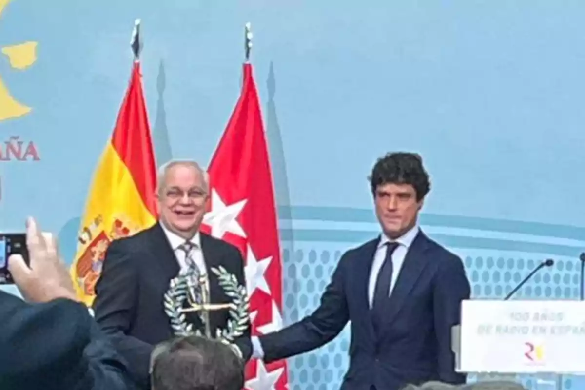 Dos hombres en un evento con banderas de España y Madrid de fondo uno de ellos sostiene un trofeo.