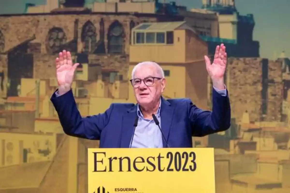 Hombre de cabello canoso y gafas, con los brazos levantados, hablando en un podio amarillo que dice "Ernest 2023" con un fondo de edificios.
