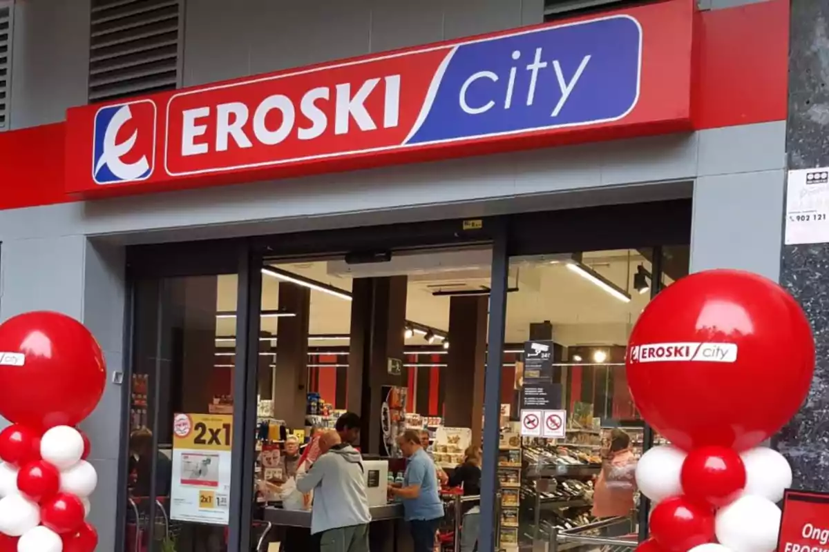 Fachada de una tienda Eroski City con globos rojos y blancos en la entrada y personas comprando en el interior.