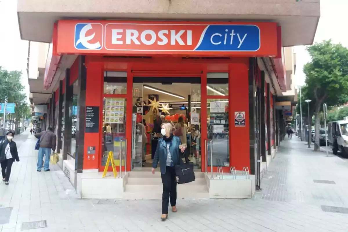 Una persona con mascarilla sale de una tienda Eroski City en una calle urbana.