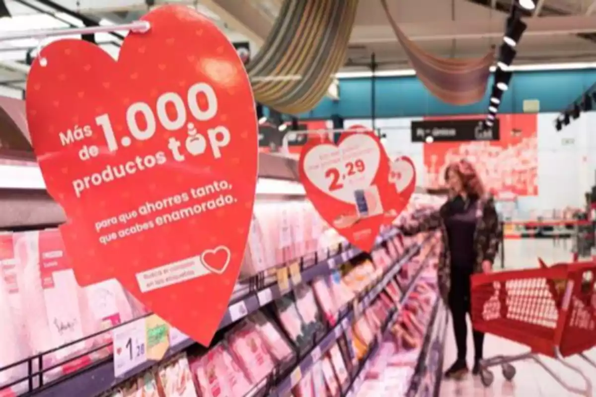 Una mujer con un carrito de compras observa productos en un supermercado, donde hay carteles en forma de corazón que anuncian ofertas y más de 1000 productos top.