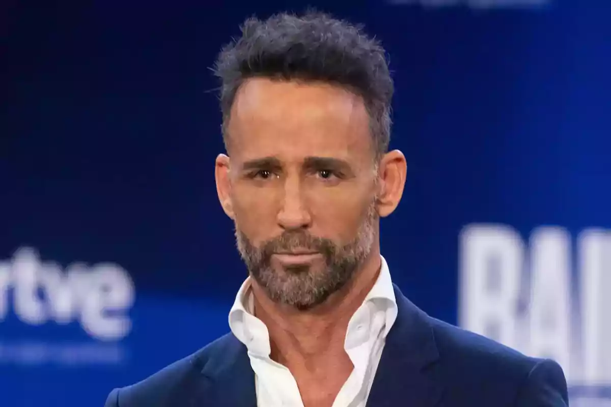 Hombre con barba y cabello corto, vestido con camisa blanca y saco oscuro, en un fondo azul.