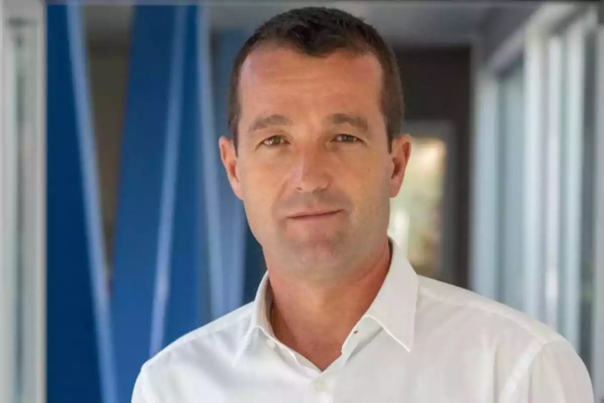 Un hombre con camisa blanca en un entorno interior con fondo azul y ventanas.