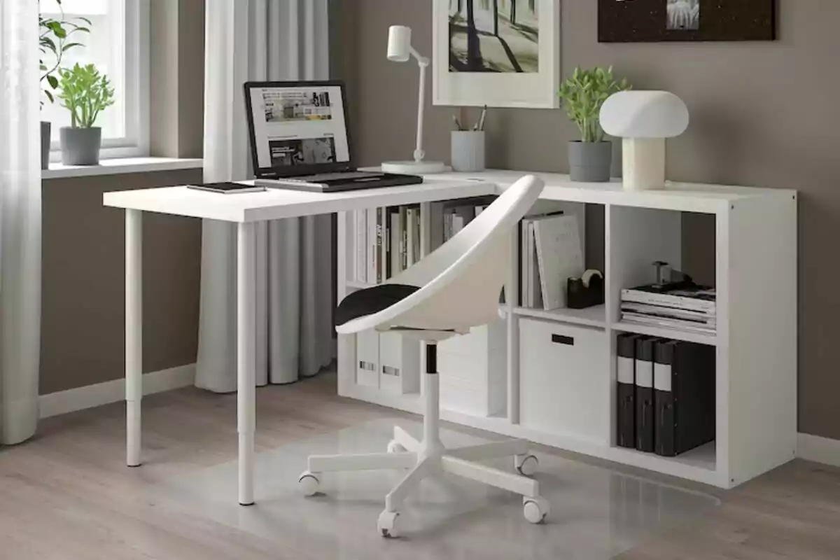 Espacio de trabajo moderno con escritorio blanco en forma de L de Ikea, silla giratoria, laptop, lámpara de escritorio y estantería con libros y plantas.