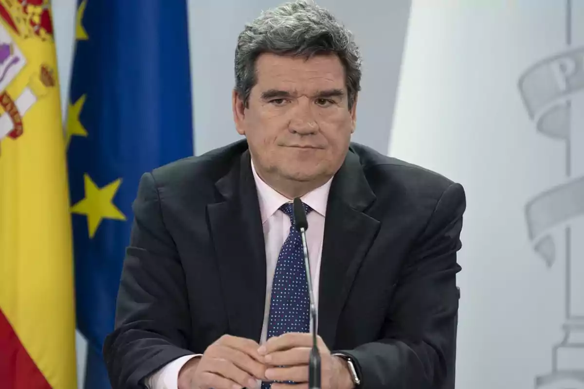 Hombre de traje oscuro y corbata azul con puntos blancos, sentado frente a un micrófono con las banderas de España y la Unión Europea en el fondo.