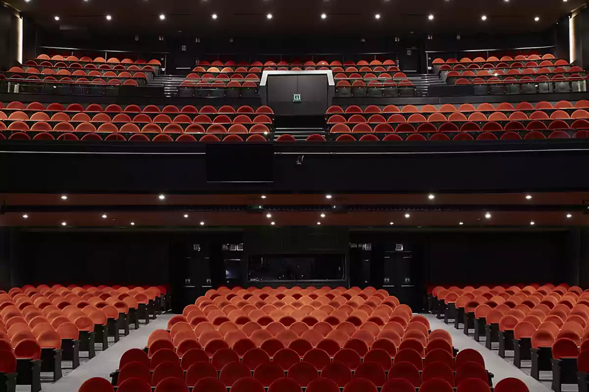 Teatro vacío con filas de asientos rojos y un balcón superior.