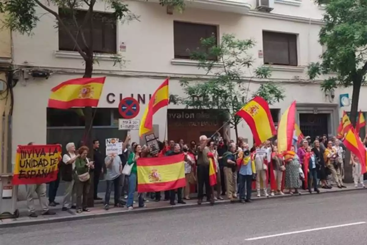 Un grupo de personas se manifiesta en la calle con banderas de España y pancartas frente a un edificio.