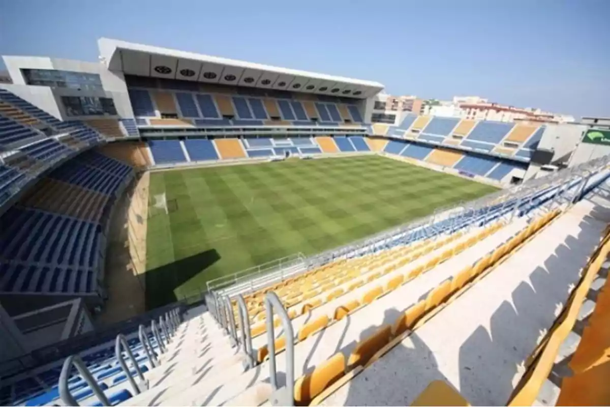 Estadio de fútbol vacío con gradas azules y amarillas y césped bien cuidado.