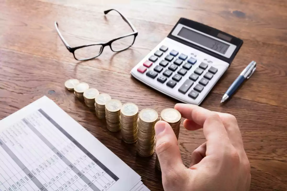 Una mano apilando monedas junto a una calculadora, un bolígrafo, unas gafas y una hoja de cálculo sobre una mesa de madera.