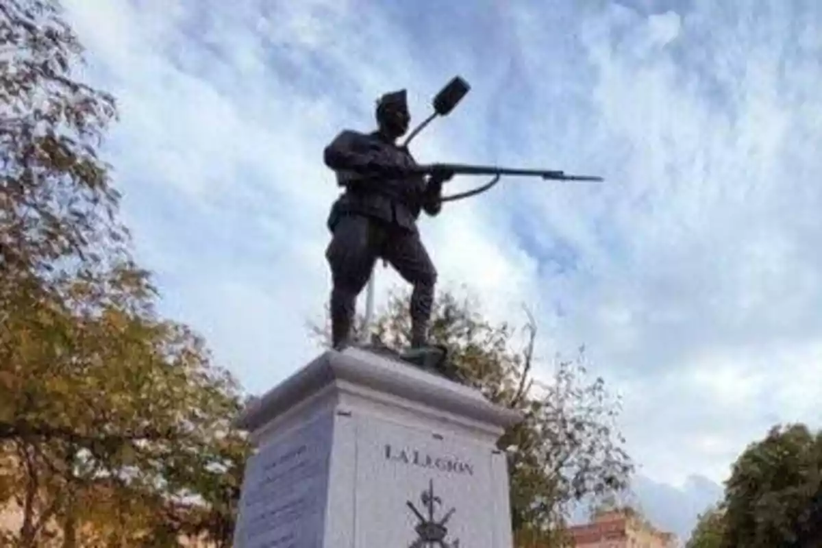 Estatua de un soldado con un arma en un pedestal con la inscripción "La Legión" y árboles en el fondo.