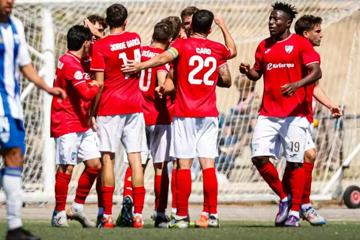 Jugadores de un equipo de fútbol con uniforme rojo celebran un gol frente a la portería mientras un rival con uniforme azul y blanco camina cerca Jugadores de un equipo de fútbol con uniforme rojo celebran un gol frente a la portería mientras un rival con uniforme azul y blanco camina cerca