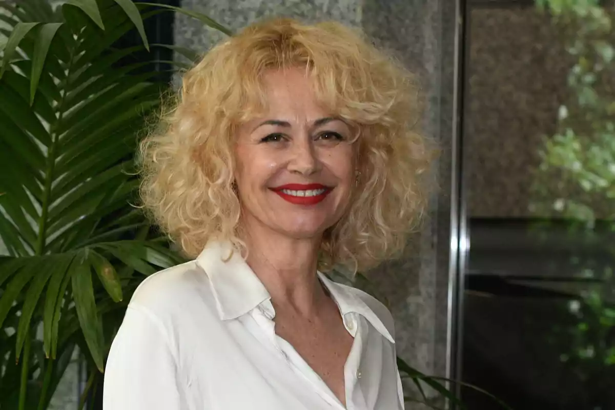 Una persona con cabello rubio rizado y camisa blanca sonriendo frente a una planta.