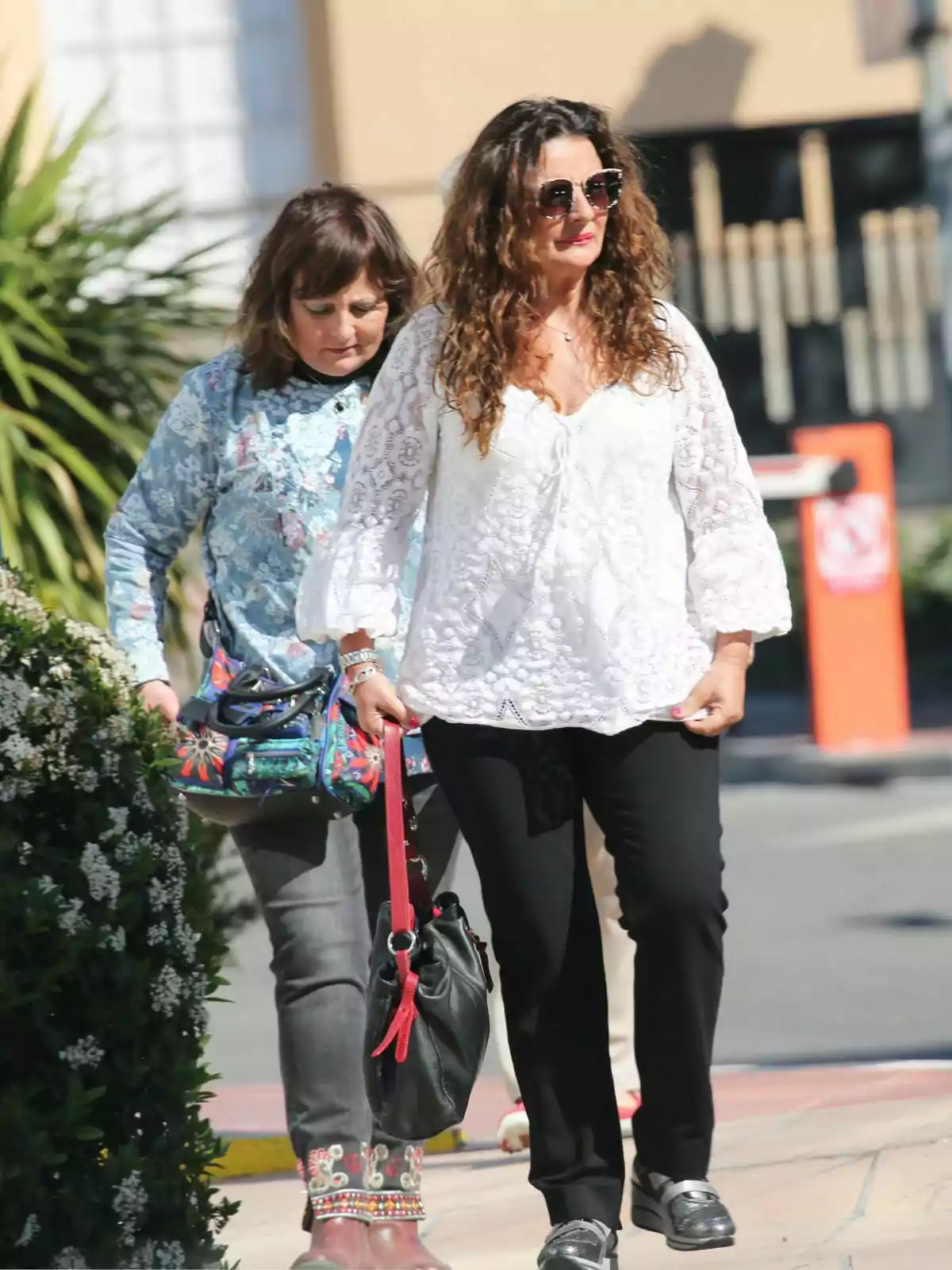 Esther Vázquez y Ana Vázquez caminando por la calle, una con blusa blanca y gafas de sol, la otra con blusa azul estampada y bolso colorido.