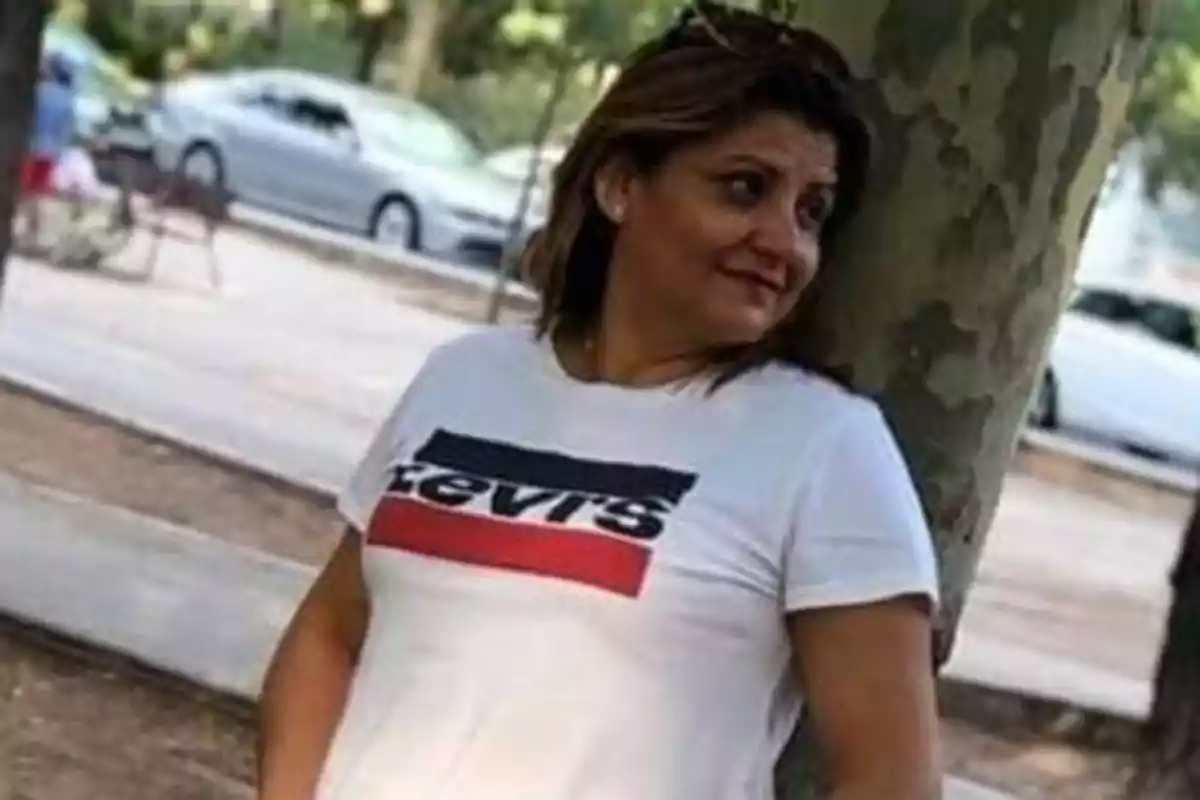 Persona con camiseta blanca apoyada en un árbol en un parque.