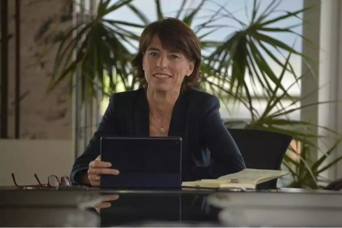 Una mujer sentada en una oficina con una tableta en las manos, un cuaderno abierto y unas gafas sobre la mesa, con plantas en el fondo.