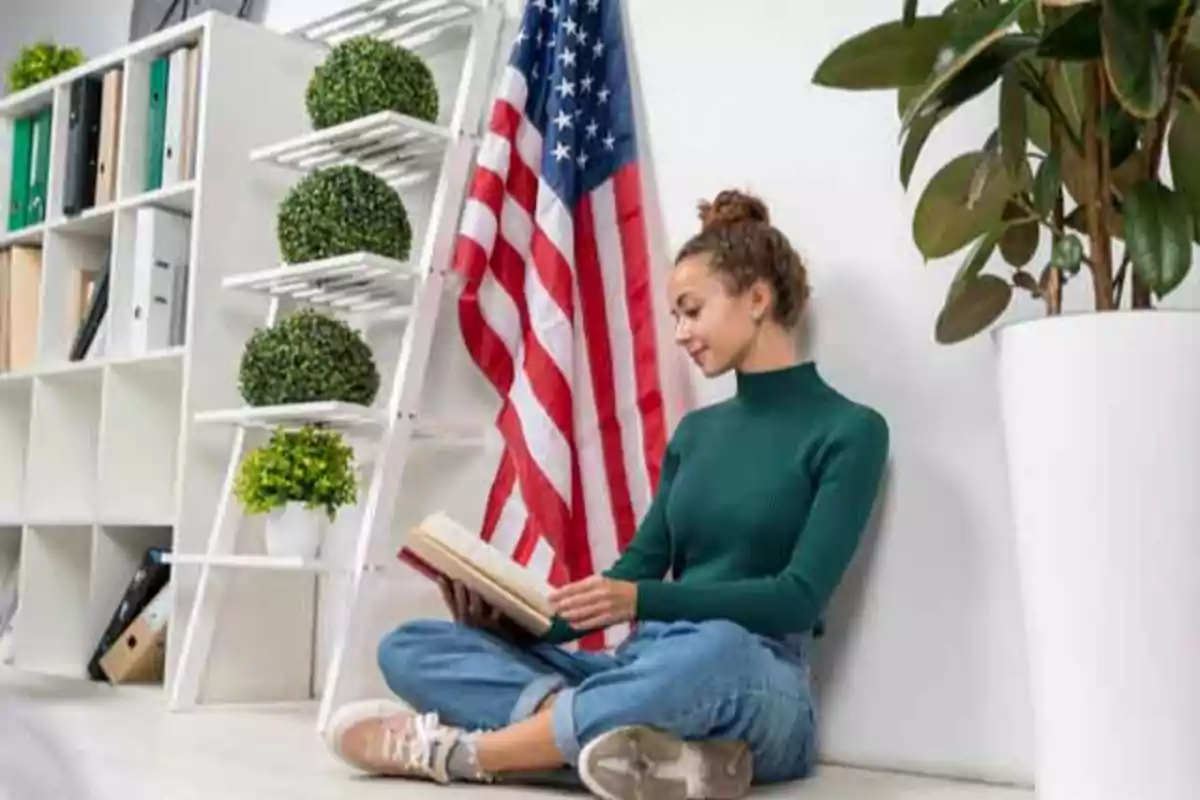 Una persona sentada en el suelo leyendo un libro junto a una bandera de Estados Unidos y una estantería con plantas.