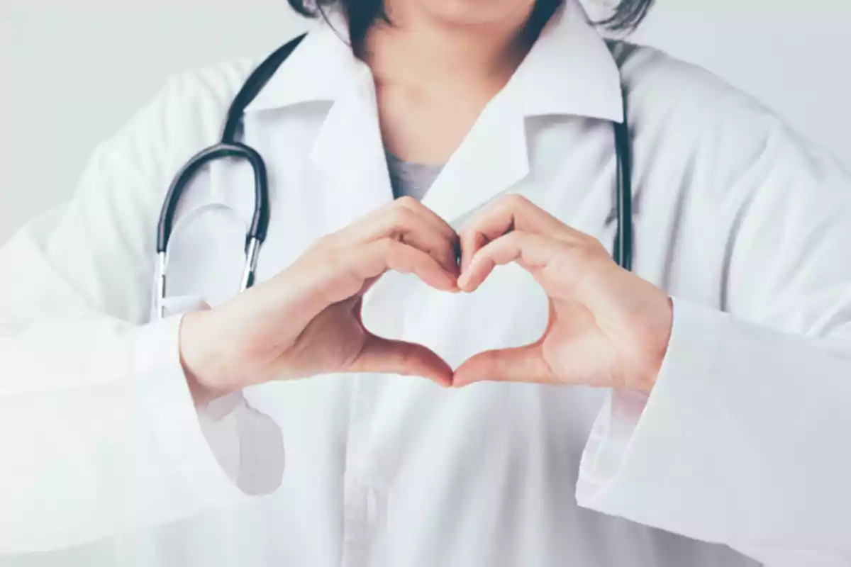 Una persona con bata médica y estetoscopio formando un corazón con las manos.