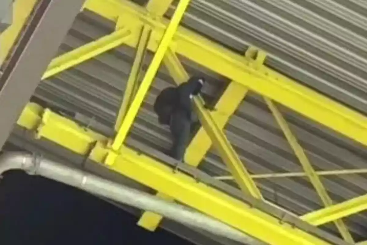 Una persona vestida de negro trepando una estructura metálica amarilla en el techo de un edificio.
