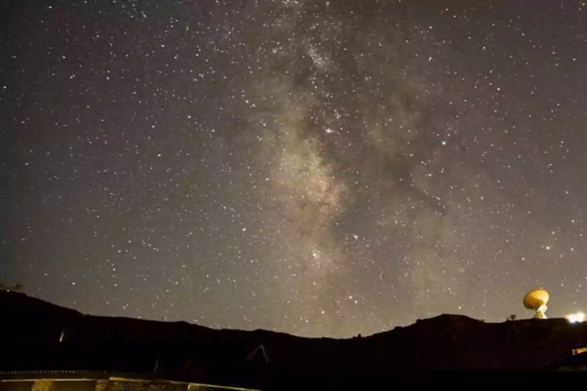 Una vista nocturna del cielo estrellado con la Vía Láctea visible y una antena parabólica en el horizonte.