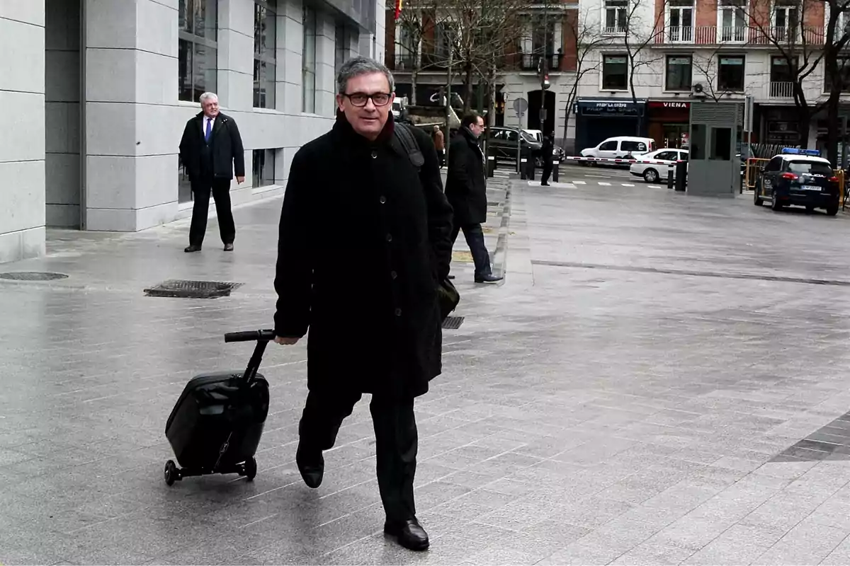 Un hombre caminando por la calle con una maleta de ruedas en un entorno urbano.