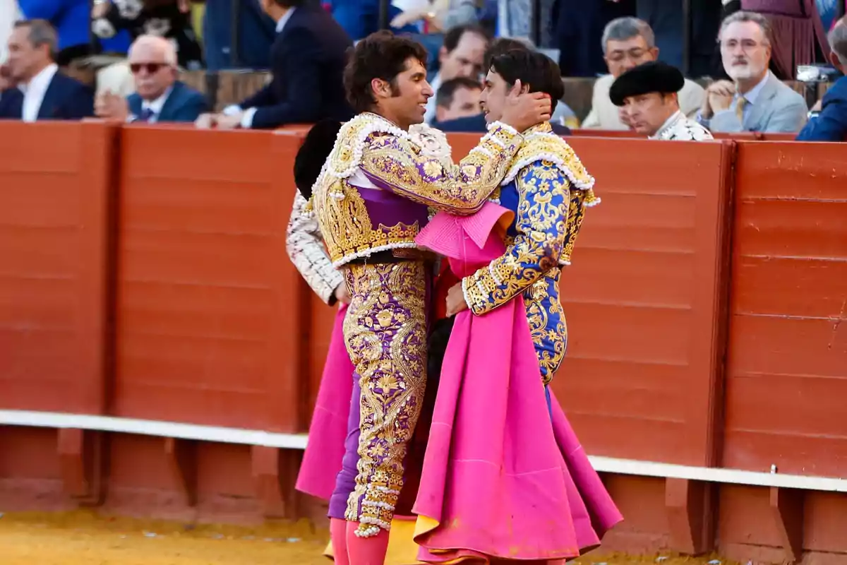 Dos toreros vestidos con trajes de luces coloridos se abrazan en una plaza de toros mientras el público observa desde las gradas.