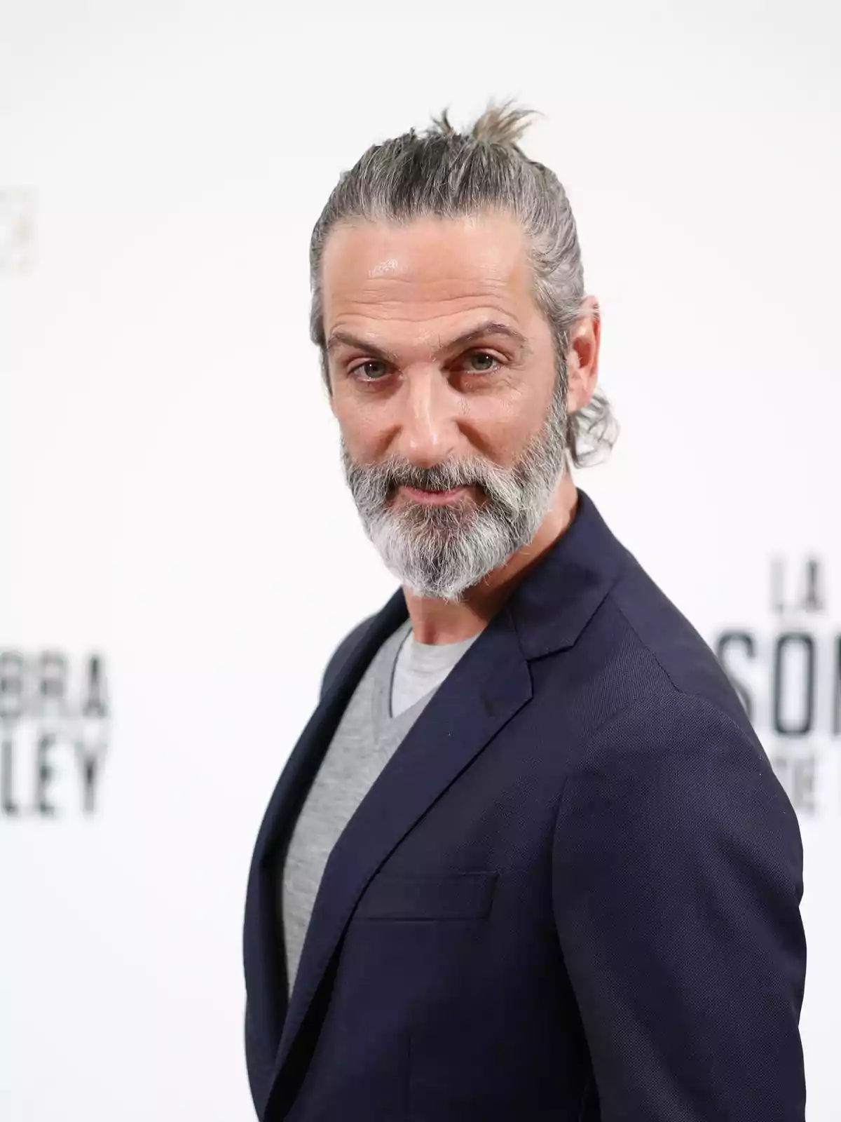 Hombre con barba y cabello recogido en un moño, vistiendo un traje oscuro y camiseta gris, posando frente a un fondo blanco. Hombre con barba y cabello recogido en un moño, vistiendo un traje oscuro y camiseta gris, posando frente a un fondo blanco.