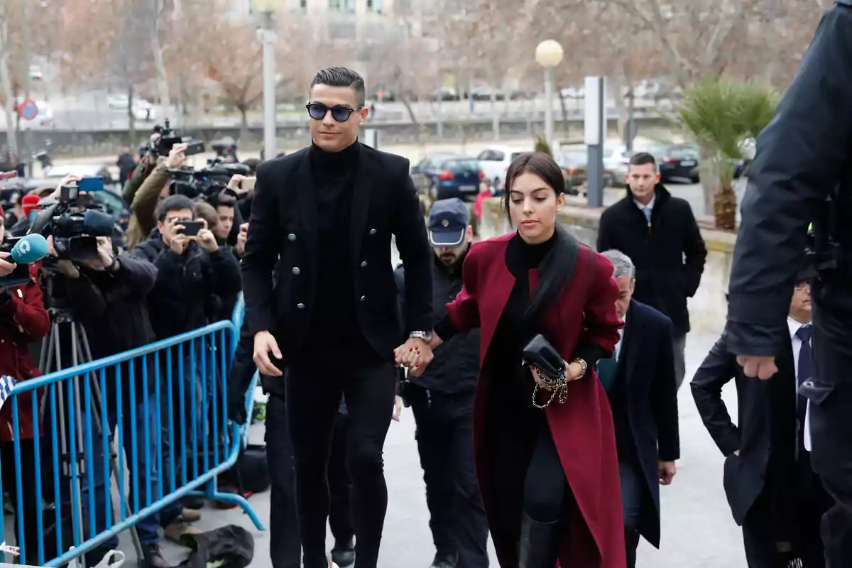 Una pareja vestida elegantemente camina de la mano mientras son fotografiados por un grupo de reporteros y camarógrafos en un evento al aire libre.