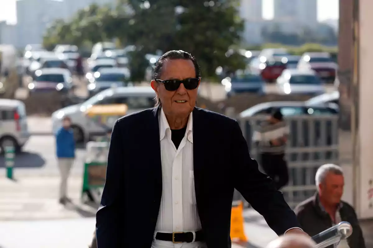 Un hombre mayor con gafas de sol y traje oscuro camina por una calle con varios coches estacionados al fondo.