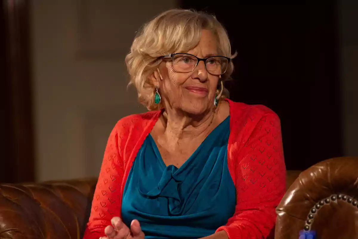 Una mujer mayor con gafas y cabello rubio, vestida con un suéter rojo y una blusa azul, sentada en un sillón de cuero.