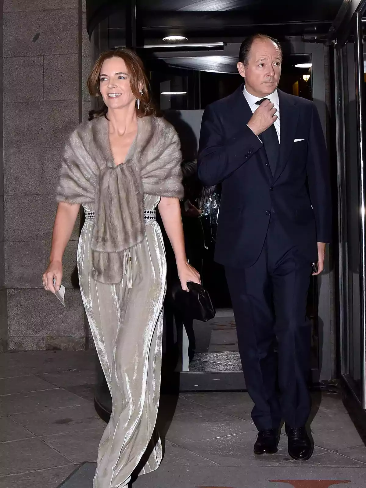 Una mujer con un elegante conjunto de pantalón y chal de piel camina junto a un hombre con traje oscuro mientras salen de un edificio Una mujer con un elegante conjunto de pantalón y chal de piel camina junto a un hombre con traje oscuro mientras salen de un edificio